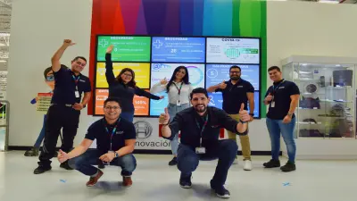 un grupo de personas se encuentran alrededor de una mesa, riendo y trabajando conjuntamente en diferentes tareas en e hackathon "Bosch Connected Experience" en Berlín. La mesa está llena de apuntes, notas coloridas, lápices y con un portátil. En el fondo, otros participantes están parados alrededor de pizarrones con pancartas de diferentes proyectos.